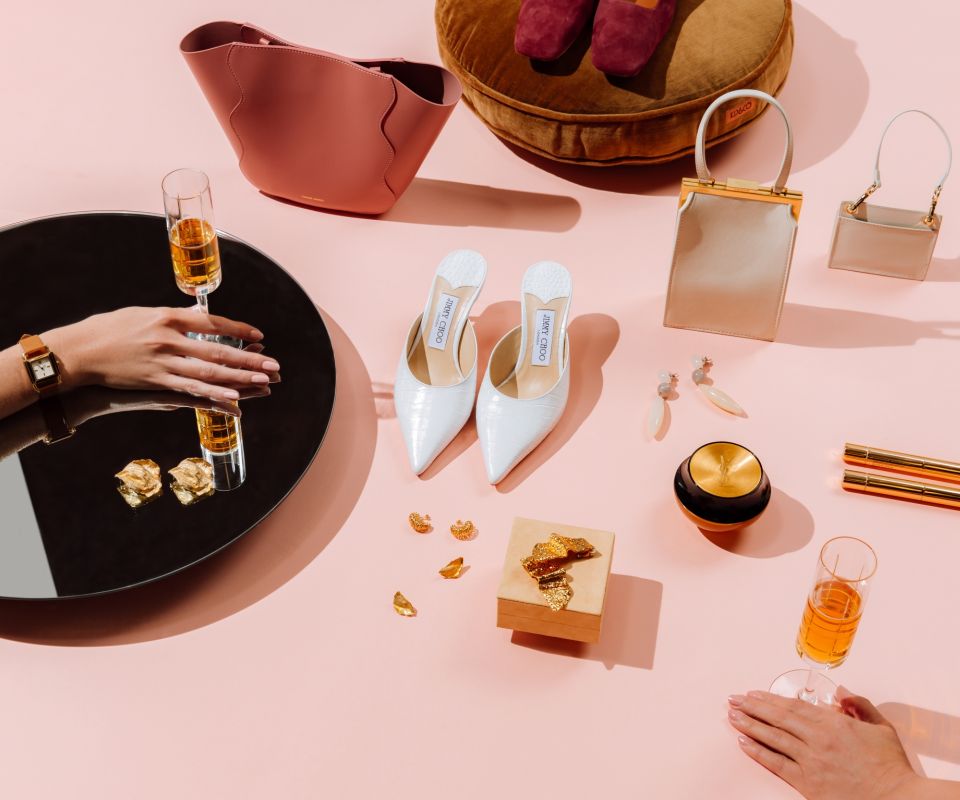 Products including shoes, handbags and glasses of wine spread over a pink surface