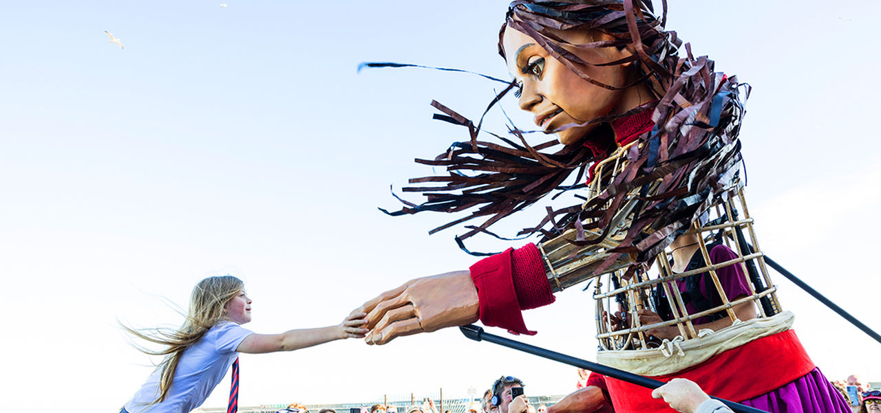 Little Amal giant puppet holding hands with a young girl