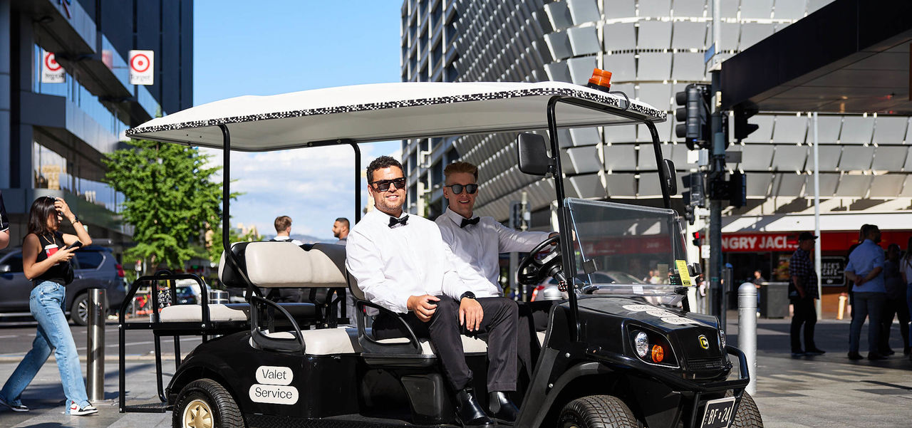 Two men dressed in bow tie, shirts, and pants, driving the Rundle Mall golf buggy through the Mall.
